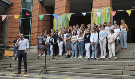 Schuldirektor Pasderski spricht ins Mikrofon. Auf den Treppnstufen dahinter stehen Schülerinnen und Schüler