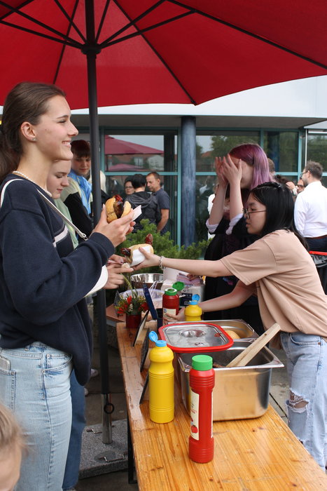 Der Grillverkaufsstand. Eine Schülerin bedient zwei Mädchen und reicht eine Wurst im Brötchen
