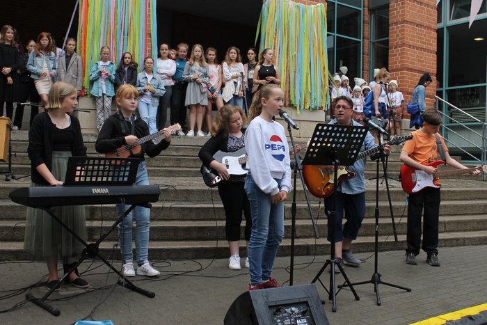 Schülerinnen und Schüler und ein Mann spielen Instrumente. Eine Schülerin singt