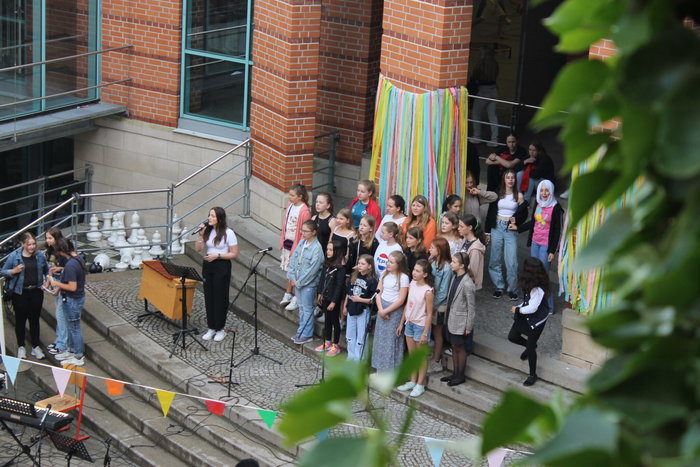 Eine Lehrerin steht vor der Schule und hält ein Mikrofon in der Hand. Hinter ihr stehen Schülerinnen