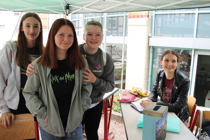 Drei Schülerinnen stehen vor einem und eine dunkelhaarige Frau sitzt auf der anderen Tischseite auf einem Stuhl