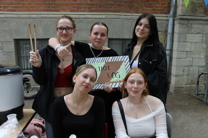 Fünf Schülerinnen lächeln in die Kamera. Eine von ihnen hält eine Holzzange in der Hand, zwei andere Schülerinnen halten ein Schild mit der Aufschrift "HOT DOG" hoch