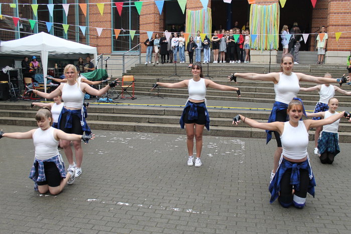 Schülerinnen in weißen Tops, schwarzen Hosen, blauer umgebundener Bluse, weißen Schuhen und schwarzen Handschuhen tanzen vor dem Schulgebäude