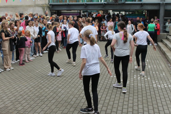 Schülerinnen mit weißen T-Shirt und schwarzen Hosen tanzen beim Hoffest. Im Hintergrund stehen viele Menschen, die zu schauen