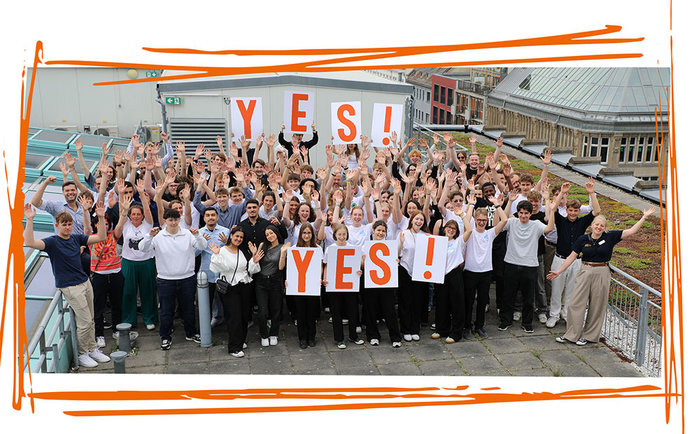 Gruppenfoto aller Teilnehmer de YES!-Wettbewerbs. Es werden Schilder mit orangenem Text hochgehalten, welcher den Namen des Wettbewerbs repräsentiert.