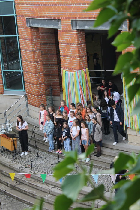 Schülerinnen stehen auf den Treppenstufen vor der Schule und eine Lehrerin hält ein Mikrofon in der Hand