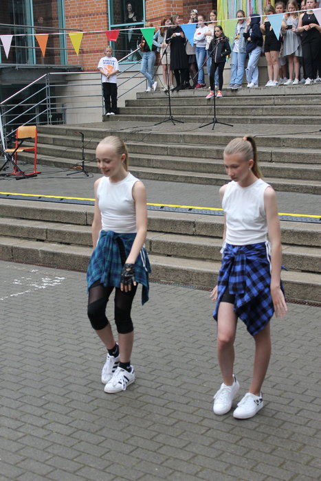 Zwei blonde Schülerrinnen in weißen Tops, schwarzen Hosen und weißen Schuhen tanzen vor dem Schulgebäude