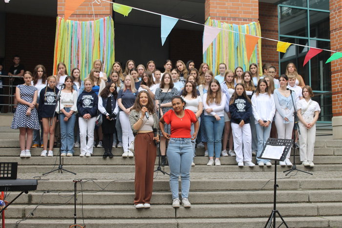 Zwei Schülerinnen stehen vor weiteren Schülerrinen und Schülern auf Treppenstufen. Eine von ihnen spricht in ein Mikrofon