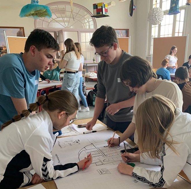Schülerinnen und Schüler arbeiten in Gruppen an einem Plakat und zeichnen das Schulgebäude